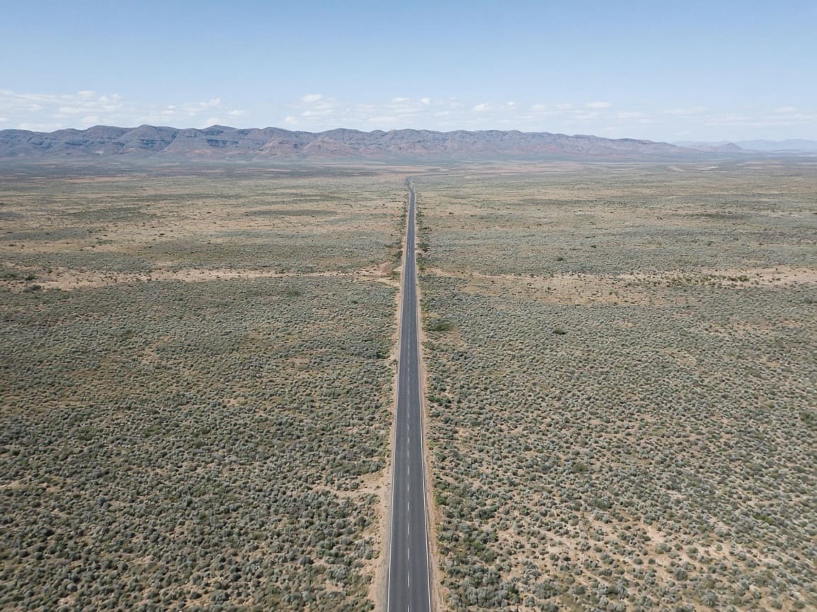 Sealed highway crossing pale plains toward distant ranges in hard Australian light